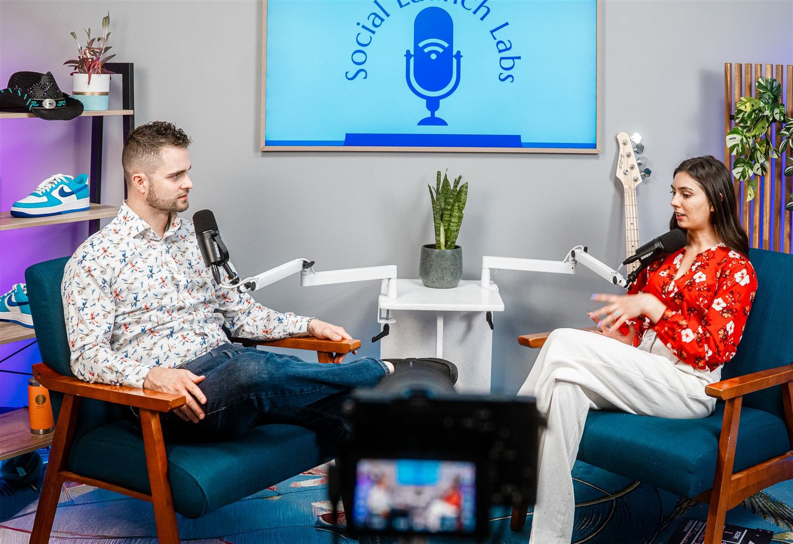 Two guests in the studio's matching teal armchairs, mid-interview with boom mics and the branded screen behind them
